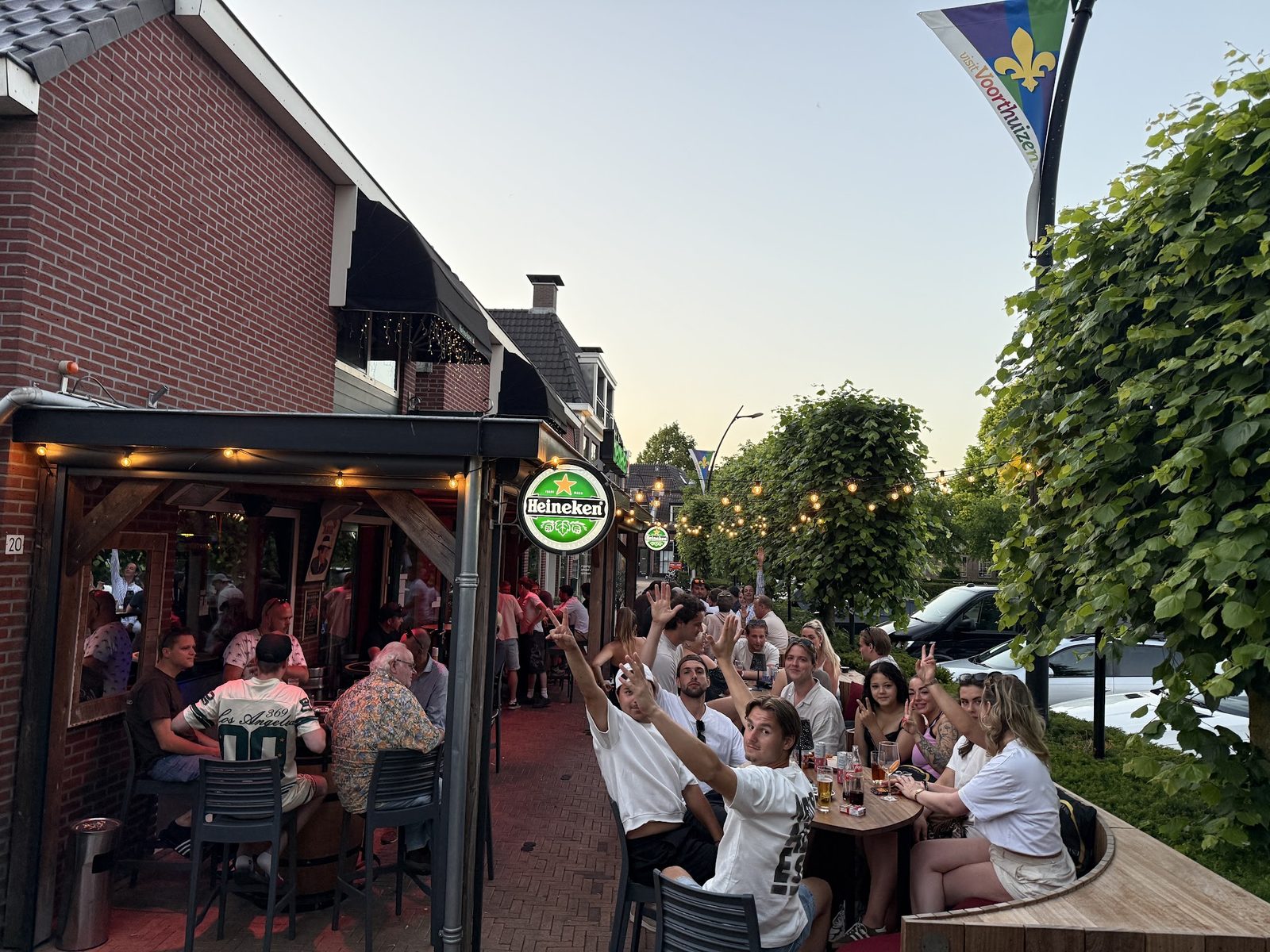 Het volle terras van Ons Kroegie aan het Smidsplein, met gasten die naar de camera zwaaien